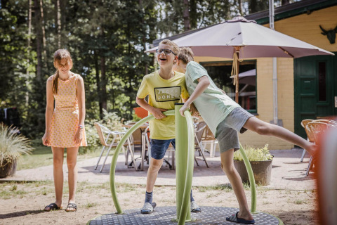 Tre børn leger sammen udenfor ved et chalet med legeredskaber, stole og parasol i sommervejret.