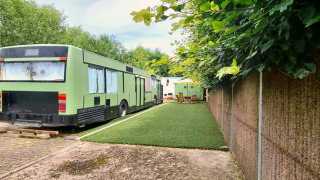 Casa móvil REEBUS verde, convertida de autobús, estacionada sobre césped artificial junto a muebles de jardín.