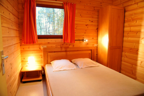 Træbeklædt soveværelse i De Boshut chalet i Belgien med dobbeltseng, lampe og udsigt til skov.