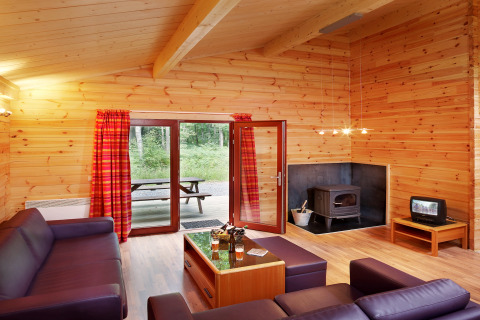 Stue i chalet De Boshut med trævægge, brændeovn, lilla sofaer og udsigt til skov, Belgien.