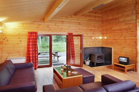 Salon du chalet De Boshut en Belgique avec murs en bois, poêle, canapés violets et vue sur la forêt.