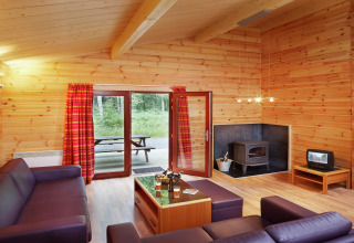 Salon du chalet De Boshut en Belgique avec murs en bois, poêle, canapés violets et vue sur la forêt.