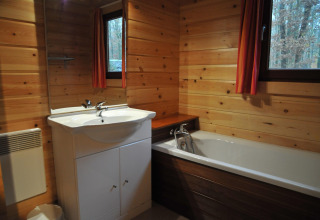 Salle de bain au chalet De Boshut en Belgique avec murs en bois, baignoire et fenêtres sur la forêt.