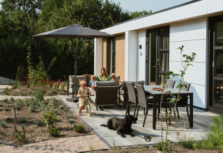 Gezin ontspant op het terras van een lodge met kinderen, hond, tuinmeubelen en een parasol.