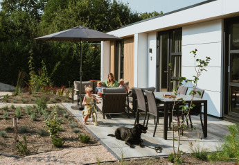 Familie entspannt sich auf der Terrasse einer Lodge mit Kindern, Hund und Garten unter einem Sonnenschirm.