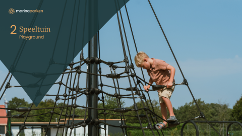Jongen klimt op touwnet in speeltuin bij glamping, onder blauwe lucht met bomen op de achtergrond.