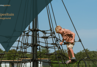 Jongen klimt op touwnet in speeltuin bij glamping, onder blauwe lucht met bomen op de achtergrond.
