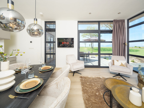 Salon moderne d'un lodge avec table à manger, TV diffusant Netflix, fauteuils confortables et vue sur des champs verts.