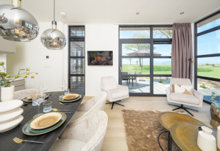 Salon moderne d'un lodge avec table à manger, TV diffusant Netflix, fauteuils confortables et vue sur des champs verts.