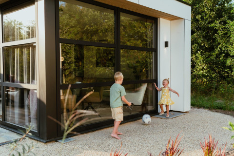 Twee kinderen spelen met een voetbal voor een modern lodge met grote ramen en omringd door groen.