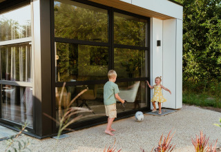 Twee kinderen spelen met een voetbal voor een modern lodge met grote ramen en omringd door groen.