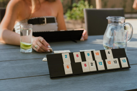 Een persoon speelt Rummikub aan een buitentafel bij een lodge, met een glas water en limoen naast zich.