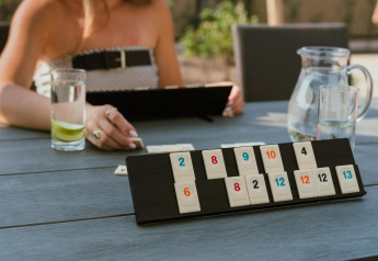 Eine Person spielt draußen an einem Tisch im Lodge-Stil Rummikub, mit Wasserkrug und Glas mit Limette.