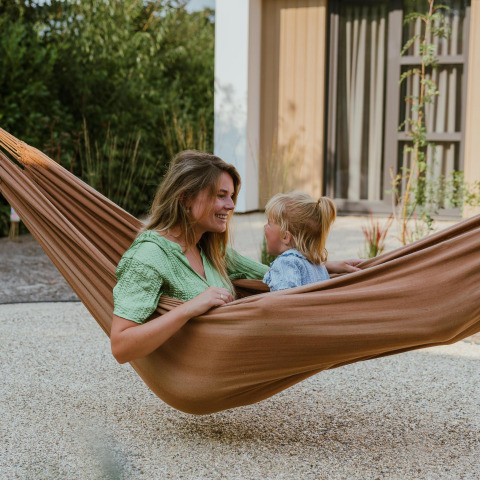 Een vrouw en een kind ontspannen samen in een bruine hangmat bij een moderne lodge met groene tuin.