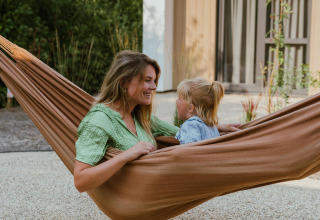 Een vrouw en een kind ontspannen samen in een bruine hangmat bij een moderne lodge met groene tuin.