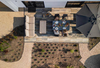 Luchtfoto van gezin dat samen eet op modern terras bij lodge, met grote parasol en tuinmeubilair.