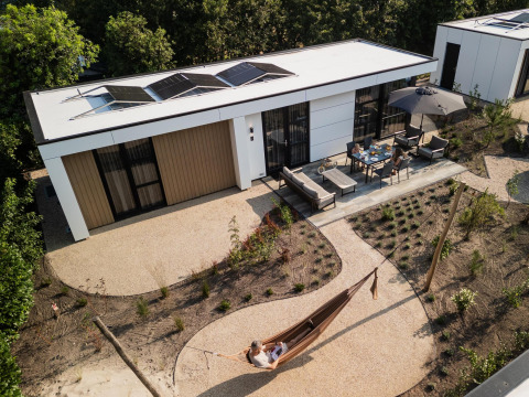 Luchtfoto van een moderne lodge met zonnepanelen, terrasmeubilair en een persoon in een tuin hangmat.