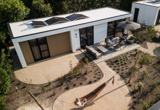 Luchtfoto van een moderne lodge met zonnepanelen, terrasmeubilair en een persoon in een tuin hangmat.