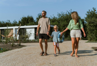 Gezin van drie loopt hand in hand voor een lodge, het kind draagt een zwemband op een zonnige dag.