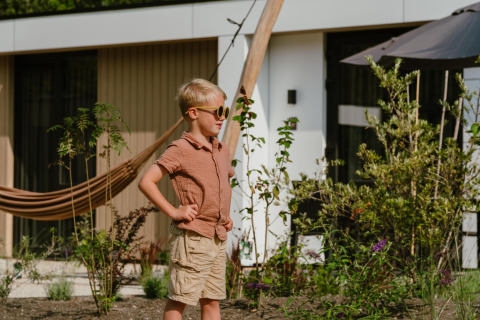 Een jongen met zonnebril en zomerkleding staat buiten bij een moderne lodge, omringd door planten.