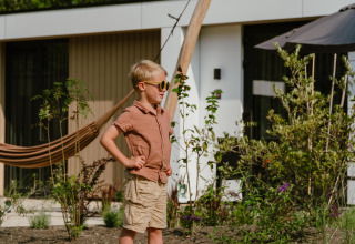 Een jongen met zonnebril en zomerkleding staat buiten bij een moderne lodge, omringd door planten.