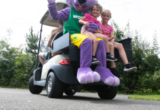 Kinderen zitten lachend naast een paarse mascotte in een golfkarretje in vakantiepark 't Rheezerwold.