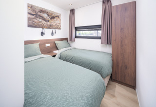 Chambre de lodge moderne avec deux lits simples, literie verte, tableau mural et fenêtre avec rideaux.