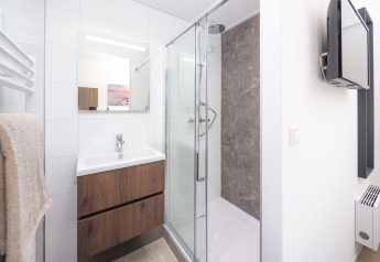 Baño moderno en un lodge con ducha de cristal, mueble de madera, espejo y televisor en la pared.