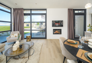 Modernes Lodge-Wohnzimmer mit großen Fenstern, Esstisch, Kamin und Blick auf eine grüne Terrasse.