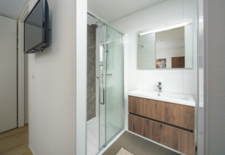 Salle de bain moderne de lodge avec douche vitrée, TV murale, meuble en bois et miroir lumineux.