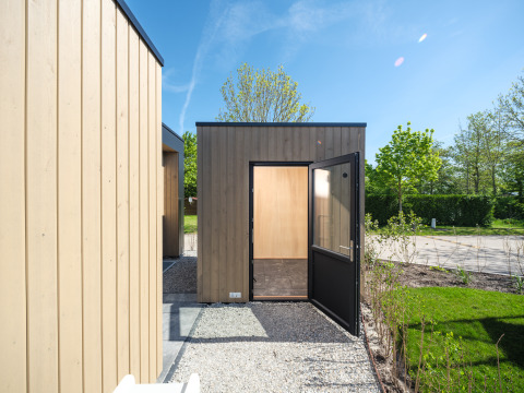 Photo d’un lodge moderne en bois avec porte ouverte, allée de gravier et verdure sous un ciel bleu clair.