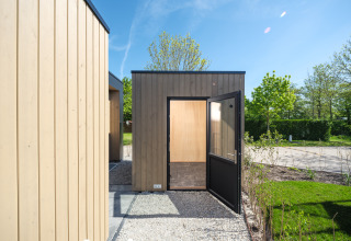 Foto di un moderno lodge in legno con porta aperta, vialetto di ghiaia e verde sotto un cielo sereno.