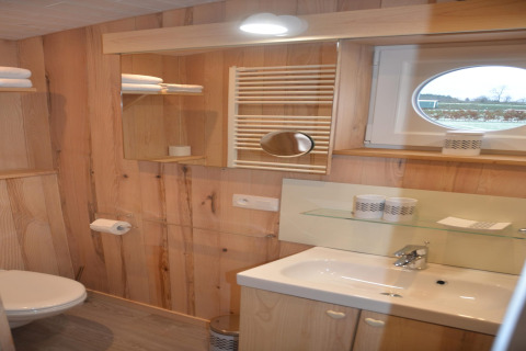 Bagno con pannelli in legno, lavandino, specchio, asciugamani e finestra rotonda a La roulotte des Ardennes, Belgio.