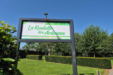 Cartello del lodge La Roulotte des Ardennes in Belgio, circondato da siepi verdi e cielo azzurro.