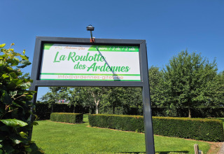 Cartello del lodge La Roulotte des Ardennes in Belgio, circondato da siepi verdi e cielo azzurro.