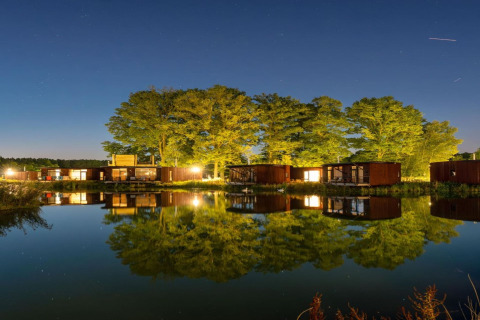 Nachtelijke glamping: moderne huisjes en bomen spiegelen in het kalme water onder een heldere sterrenhemel.