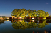 Night view of glamping site with modern cabins and trees reflecting in calm water under a starry sky.
