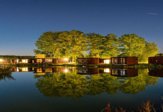 Nachtelijke glamping: moderne huisjes en bomen spiegelen in het kalme water onder een heldere sterrenhemel.