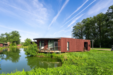 Moderne houten lodge aan het water, omringd door groen en bomen onder een heldere blauwe lucht.
