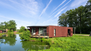 Moderne houten lodge aan het water, omringd door groen en bomen onder een heldere blauwe lucht.