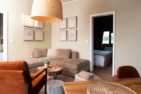 Stijlvolle glampingaccommodatie met comfortabele hoekbank, oranje stoelen en zicht op de slaapkamer.