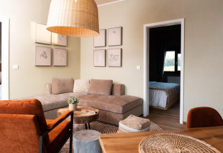 Stijlvolle glampingaccommodatie met comfortabele hoekbank, oranje stoelen en zicht op de slaapkamer.