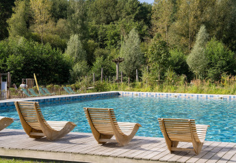 Scenic outdoor swimming pool with unique wooden loungers, nestled in lush greenery at a glamping site.