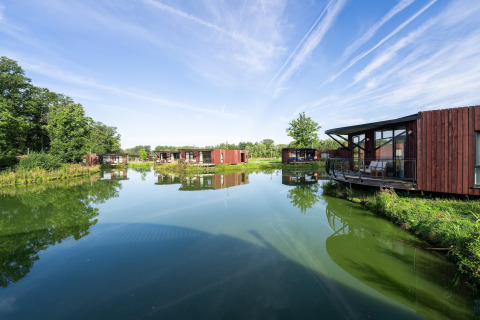 Moderne Lodges am ruhigen See, umgeben von grüner Natur und blauem Himmel mit weißen Streifenwolken.