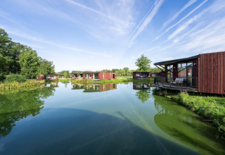 Modern lodge cabins by a tranquil lake, surrounded by lush greenery and blue sky with contrail streaks.