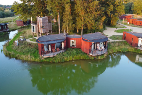 Luchtfoto van moderne rode lodges aan een meer, omringd door groene bomen, paden en rustige natuur.