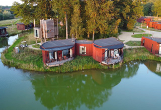 Luchtfoto van moderne rode lodges aan een meer, omringd door groene bomen, paden en rustige natuur.