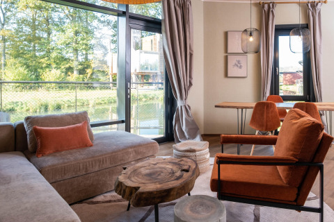 Moderne glamping accommodatie met grote ramen, comfortabele zitplaatsen en houten tafels in warme tinten.