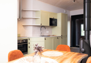 Moderne glampingkeuken met groene kasten, houten eettafel en oranje stoelen in een lichte ruimte.