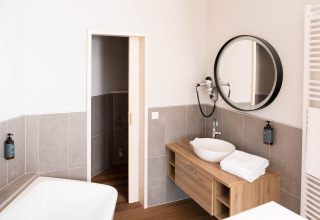 Moderne badkamer in glamping-accommodatie met ronde spiegel, houten wastafel en stijlvolle badkuip.